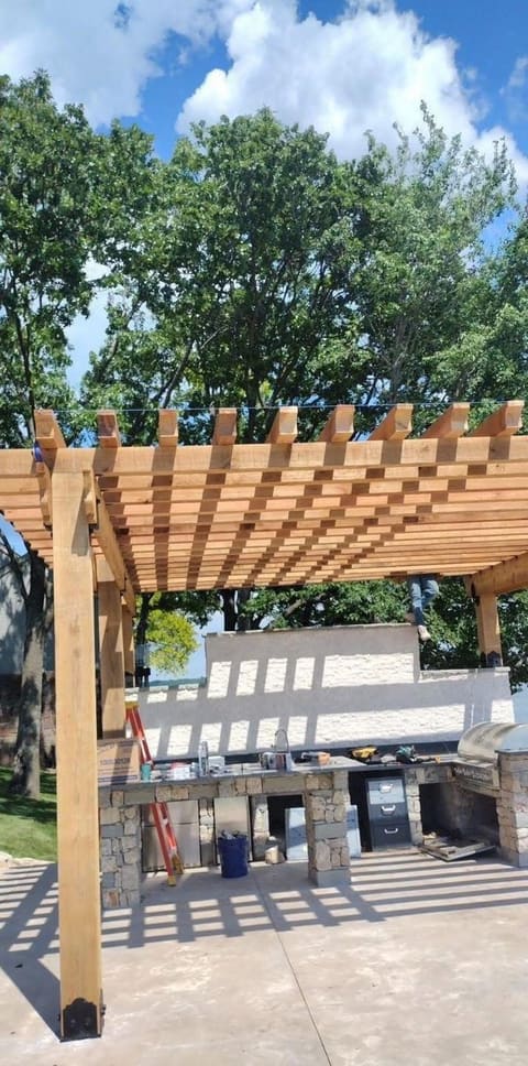 Custom cedar pergola built over a stone outdoor kitchen and bar under a blue summer sky