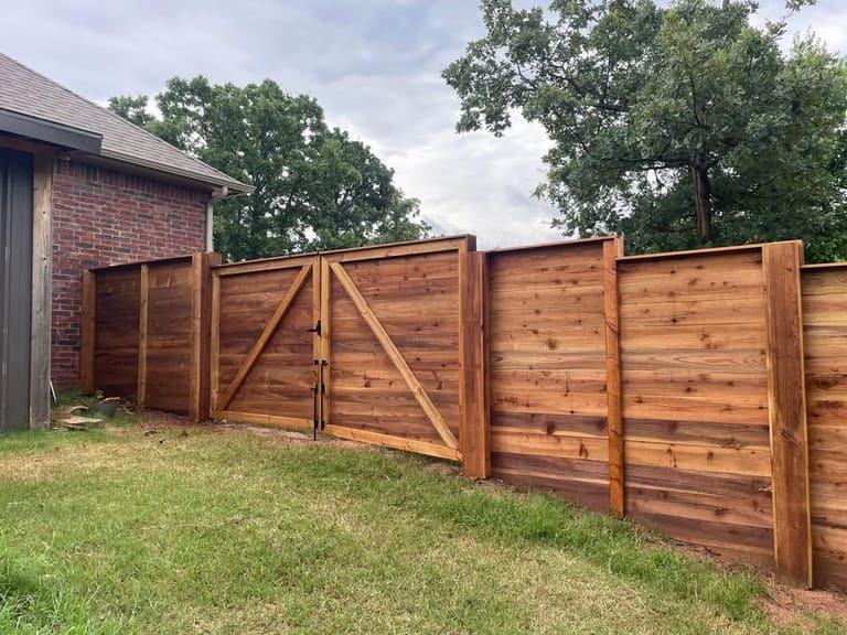 Newly built cedar privacy fence with a double swing gate and black hardware running alongside a brick home