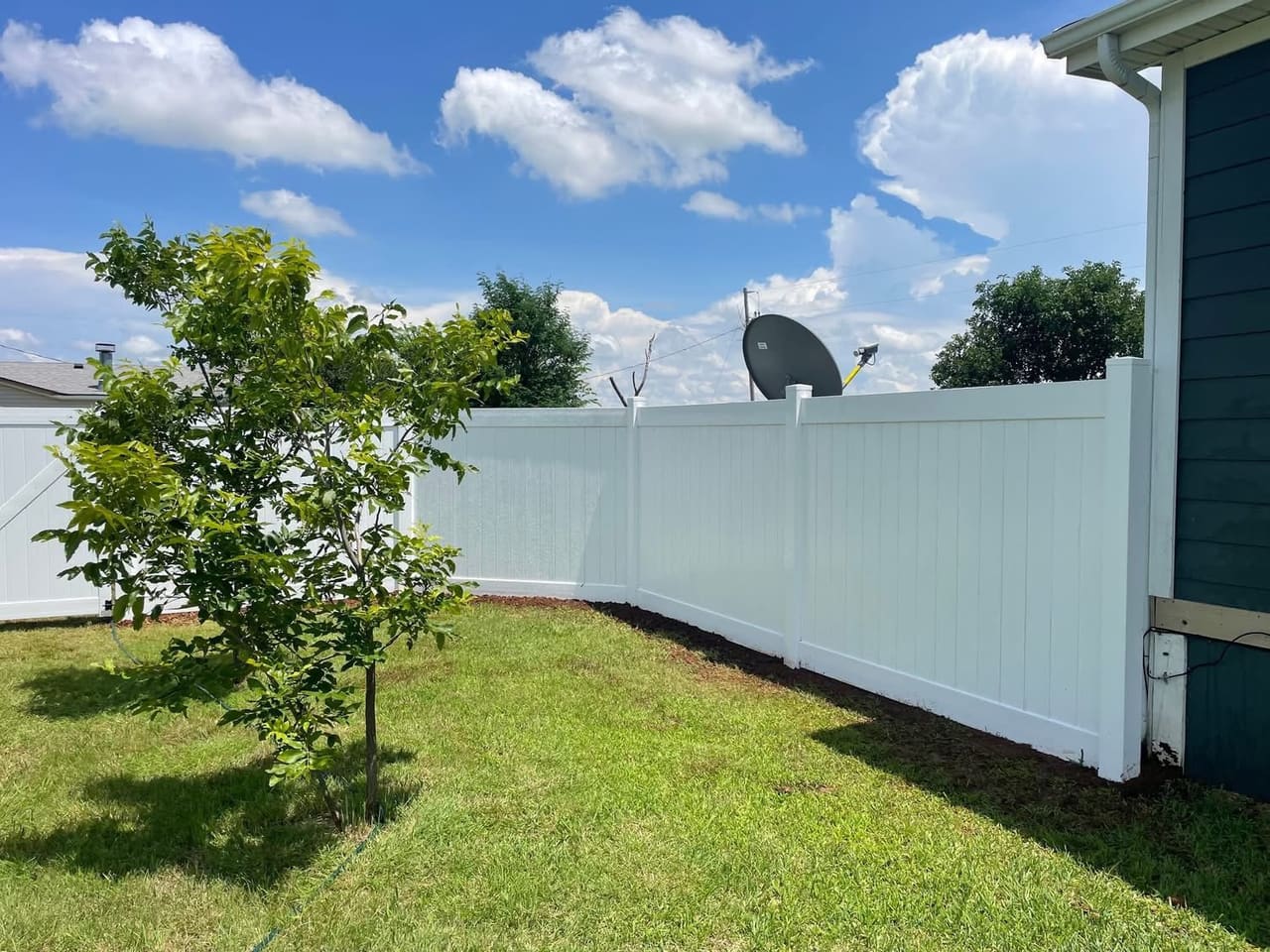 White vinyl privacy fence enclosing a green backyard beside a home under a blue Oklahoma sky